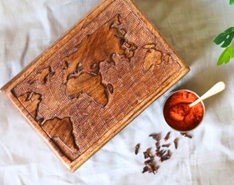May include: A rectangular wooden serving tray with a carved world map design. The tray is brown and has a textured surface. A small silver bowl with red powder and a spoon sits next to the tray. Scattered cloves are also present.