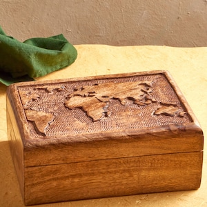 May include: A rectangular wooden box with a carved world map design on the lid. The box is made of light brown wood and has a textured surface. A green cloth is visible in the background.