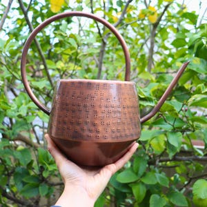 May include: A copper-coloured metal watering can featuring a wide, rounded body with a textured pattern of small, recessed square indentations. It includes a tall, arched handle and a long, slender spout extending from the side of the vessel.