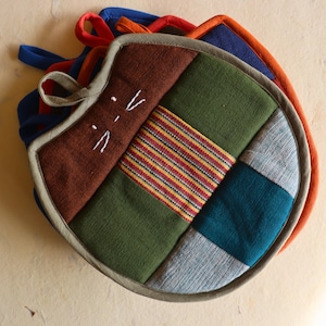 May include: A collection of cat-shaped quilted pot holders in various colours. The main pot holder has a brown face with white embroidered whiskers, and a patchwork design of green, teal, and striped fabric. Other pot holders in blue and orange are visible.