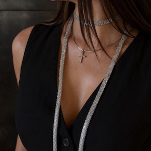 Puede incluir: Un collar de cadena de malla plateada usado como un accesorio largo tipo bufanda, combinado con una gargantilla plateada y un delicado collar con colgante de cruz plateada con pequeñas piedras incrustadas, sobre una blusa negra sin mangas.