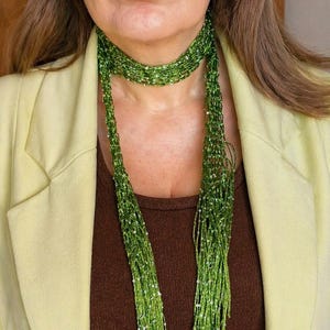 May include: A long, thin, green scarf constructed from a delicate, open-knit mesh material. The scarf is embellished with numerous small, reflective silver sequins scattered throughout the fabric, creating a shimmering effect against the green yarn.