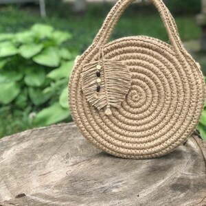 May include: A round, tan crocheted bag with a woven handle. The bag has a small, tan crocheted feather charm attached to it.