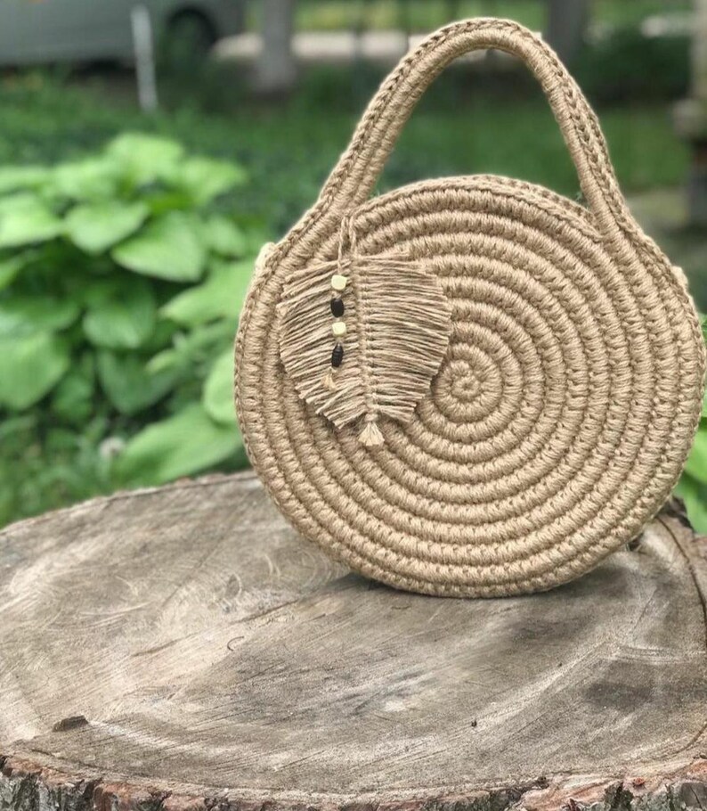 May include: A round, tan crocheted bag with a woven handle. The bag has a small, tan crocheted feather charm attached to it.