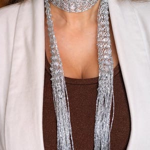 May include: A silver woven necklace with long, dangling strands. The necklace is worn over a brown top and a white blazer. The necklace has a delicate, shimmering appearance, suitable for formal or casual wear.