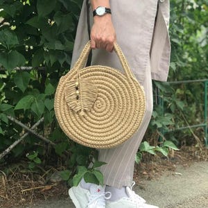 May include: A round, woven straw bag with a brown leather handle and a small, decorative tassel. The bag is made of natural materials and has a rustic, bohemian style.