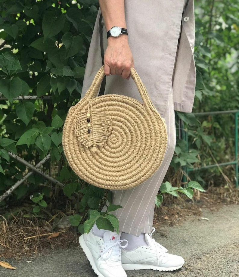 May include: A round, woven straw bag with a brown leather handle and a small, decorative tassel. The bag is made of natural materials and has a rustic, bohemian style.
