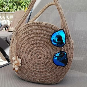 May include: A round, crocheted bag made of natural fibre with a long strap. The bag is brown and has a circular design. A pair of blue sunglasses are resting on the bag.