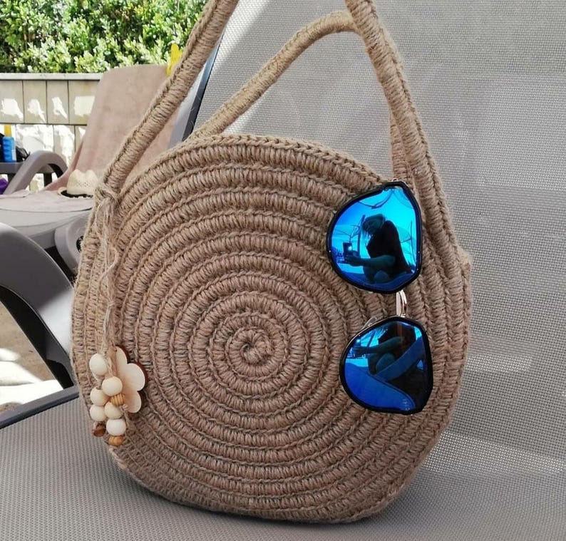 May include: A round, crocheted bag made of natural fibre with a long strap. The bag is brown and has a circular design. A pair of blue sunglasses are resting on the bag.
