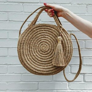 May include: A round, woven straw bag with a long strap and a tassel. The bag is light brown and has a circular shape.