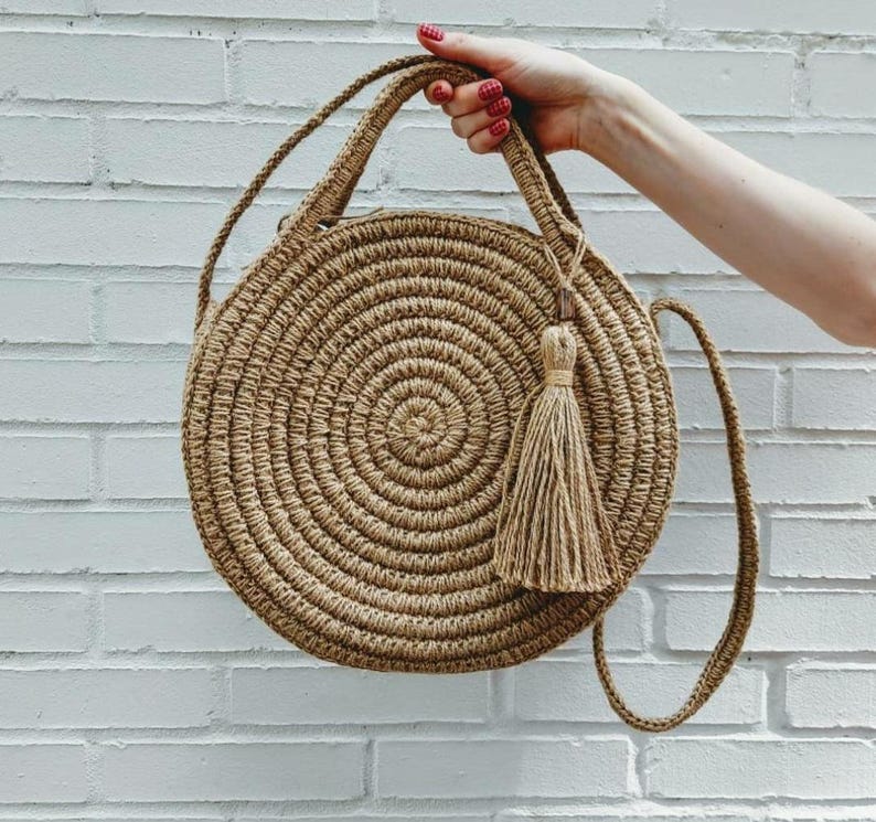 May include: A round, woven straw bag with a long strap and a tassel. The bag is light brown and has a circular shape.