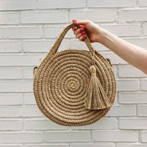 May include: A round, woven straw bag with a brown tassel. The bag has a circular shape and is made of natural materials.