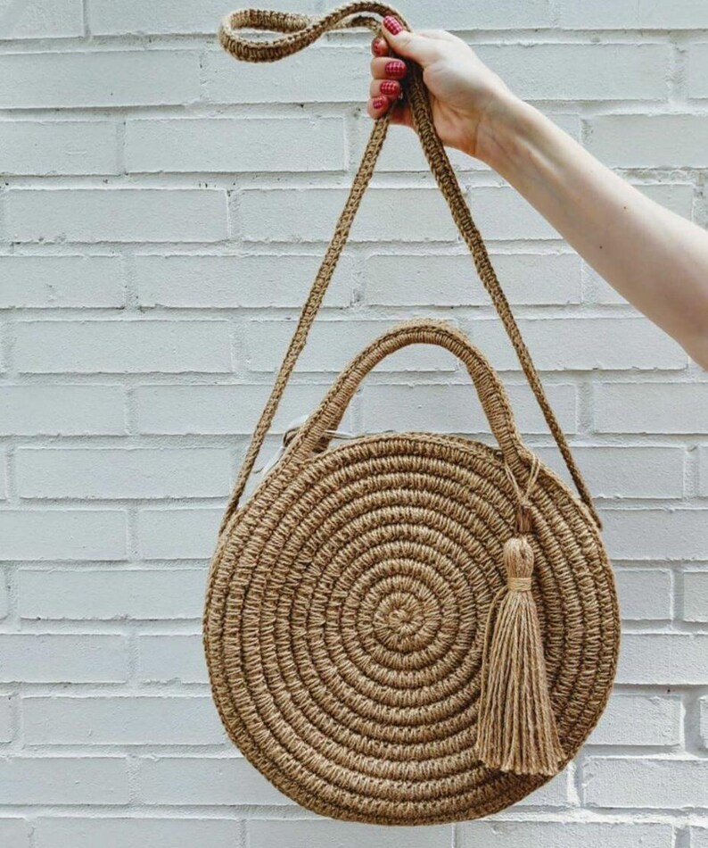 May include: A round, woven straw bag with a long strap and a tassel. The bag is brown and has a circular design.
