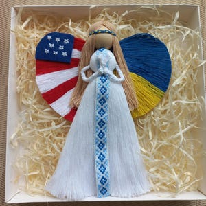 May include: A white yarn angel doll with blonde yarn hair, wearing a blue and white embroidered ribbon. The angel has wings made of yarn in the colors of the American and Ukrainian flags.