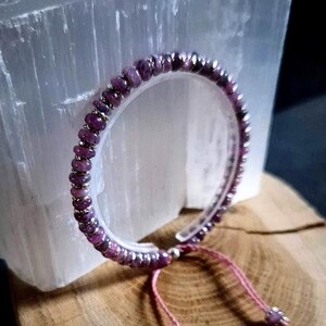 Könnte beinhalten: Ein Armband mit kleinen, runden, lila Edelsteinen und silberfarbenen Abstandshaltern. Das Armband befindet sich auf einem transparenten, flexiblen Band mit einem rosa Kordelverschluss. Es liegt auf einer Holzoberfläche mit einer weißen Kristallformation im Hintergrund.
