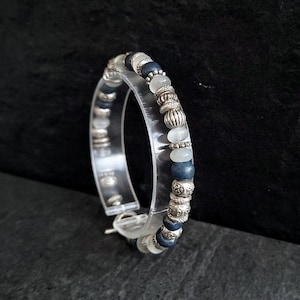 Puede incluir: Un brazalete de cuentas plateadas y azules con un cierre de plástico transparente. Las cuentas son redondas y tienen una superficie ligeramente texturizada.