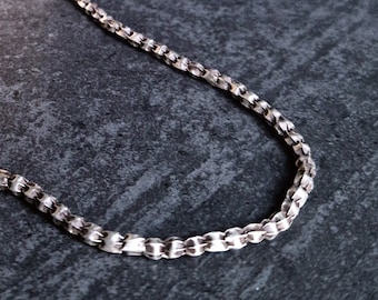 May include: A silver-colored chain necklace with a box chain design. The necklace is displayed on a wooden block against a dark gray background. The chain is made of small, rectangular links.