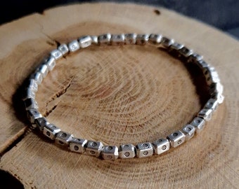 May include: A circular bracelet composed of small, metallic, cube-shaped beads. Each individual bead features a smooth, reflective surface with a tiny, centered indentation. The bracelet forms a continuous loop, showcasing a uniform, repeating pattern of these silver-toned, geometric components arranged in a simple, linked design.