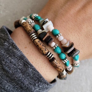 Peut inclure: Un ensemble de trois bracelets en turquoise, bois et coquillages. Les bracelets sont empilés sur le poignet d'une personne.