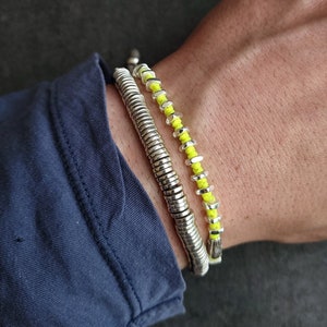 May include: Two bracelets on a wrist. One is a silver metal coil design, and the other is a beaded bracelet with yellow beads. The wrist is covered by a blue long-sleeve shirt.