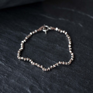 Peut inclure: Un bracelet en argent et marron avec un fermoir en argent. Le bracelet est composé de petites perles carrées enfilées sur un cordon marron.