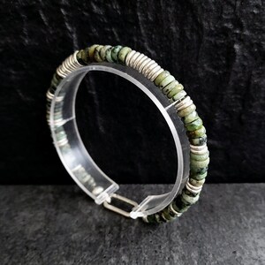 May include: A silver bracelet with green gemstone beads. The bracelet is made of small, round beads that are strung together on a silver wire. The beads are a light green color with darker green flecks.