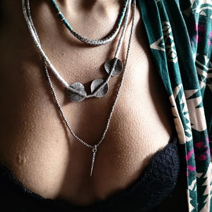 May include: A layered necklace with three silver pendant discs and a silver pendant in the shape of a spear. The necklace is worn over a black lace bra and a green and white patterned shirt.