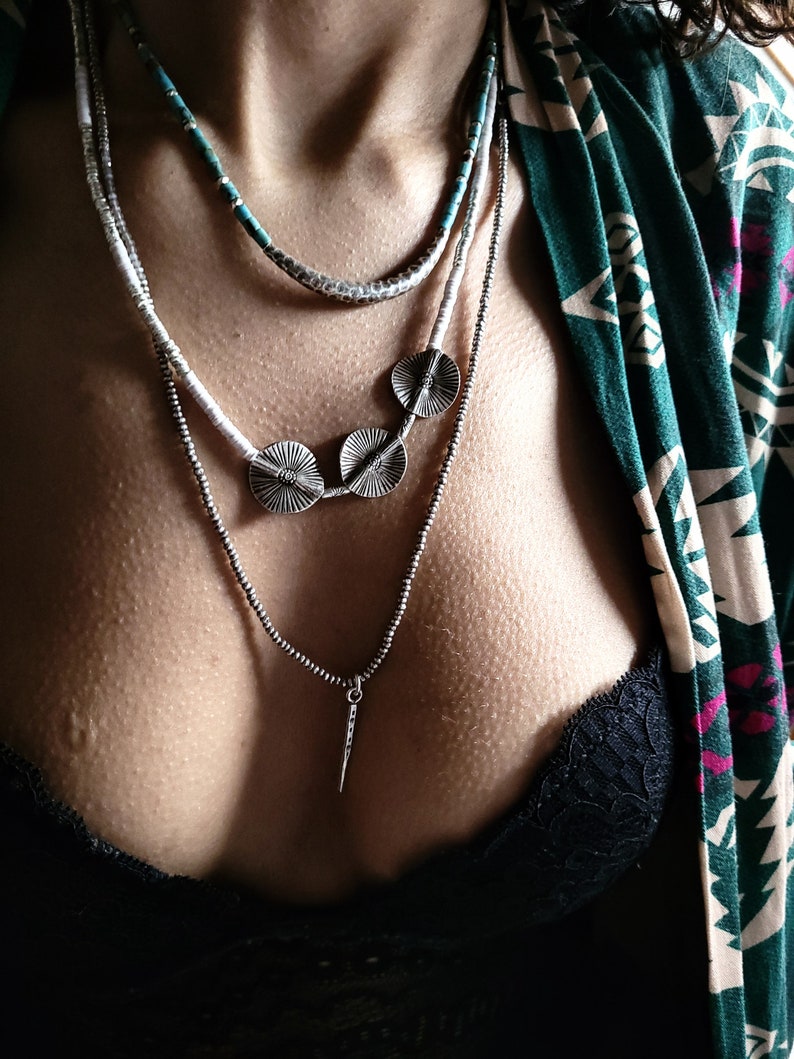 May include: A layered necklace with three silver pendant discs and a silver pendant in the shape of a spear. The necklace is worn over a black lace bra and a green and white patterned shirt.
