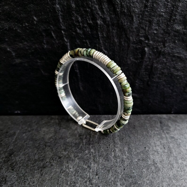 May include: A silver bracelet with green gemstone beads. The bracelet is made of small, round beads that are strung together on a silver wire. The beads are a light green color with darker green flecks.