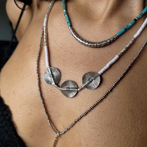 May include: Layered silver necklaces with turquoise and white beads. The necklace features three silver pendant discs with a textured design.
