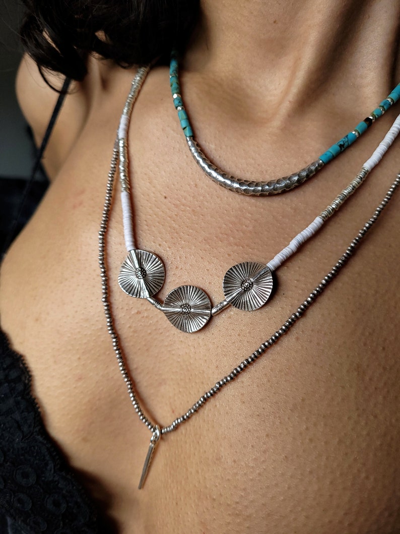 May include: Layered silver necklaces with turquoise and white beads. The necklace features three silver pendant discs with a textured design.