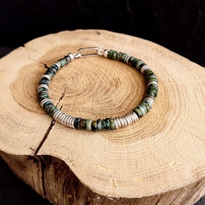 May include: A green and silver beaded bracelet with a silver clasp. The bracelet is made of small, round beads and is lying on a wooden surface.