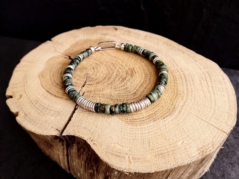 May include: A green and silver beaded bracelet with a silver clasp. The bracelet is made of small, round beads and is lying on a wooden surface.