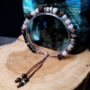 Peut inclure: Un bracelet en perles brunes et blanches avec un élastique noir et un cordon brun. Le bracelet est composé de perles en bois, de perles blanches et de perles argentées.