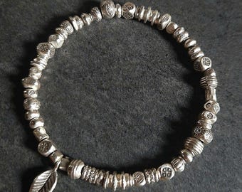 Op de afbeelding: Zilveren kralenarmband met een bladbedel. De armband heeft verschillende vormen en maten zilveren kralen, waaronder ronde, vierkante en cilindrische. De bladbedel hangt aan de armband. De armband ligt op een donkergrijze ondergrond.