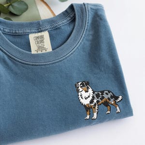 Könnte beinhalten: Blaues Jeans-farbenes Comfort Colors T-Shirt mit einem gestickten Australian Shepherd Hunde-Design. Der Hund hat schwarze, weiße und braune Markierungen. Das Shirt hat einen Rundhalsausschnitt und ein Etikett mit der Aufschrift "Comfort Colors".