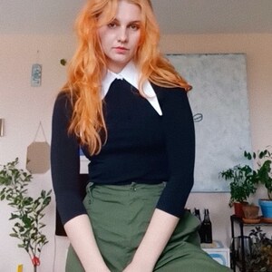 Peut inclure: Une personne porte un pull noir avec un col blanc et un pantalon vert olive. La personne a de longs cheveux roux ondul&eacute;s. L'arri&egrave;re-plan comprend des plantes en pot et une peinture.