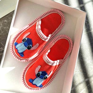 Op de afbeelding: Een paar rode pantoffels met een blauw cartoonkarakterontwerp. De pantoffels hebben een wit en roze gestreepte rand en zitten in een witte doos. Het cartoonkarakter heeft grote zwarte ogen en grote oren.