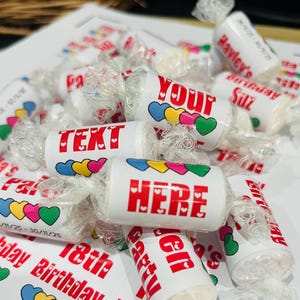 May include: A collection of wrapped sweets with red text and colourful heart designs. The text on the sweets reads "TEXT HERE" and "YOUR". Some sweets have the text "Hayley's Birthday" and dates. The sweets are white with clear wrapping.