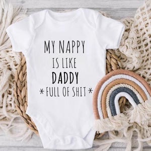 May include: White baby bodysuit with the text "MY NAPPY IS LIKE DADDY * FULL OF SHIT *" in black. A small, colourful rainbow decoration is next to the bodysuit. The bodysuit is on a woven basket.