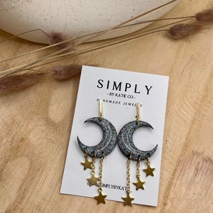 Op de afbeelding: Een paar oorbellen in de vorm van een halve maan met een donkere, glinsterende afwerking en bungelende goudkleurige sterbedels aan fijne kettinkjes, gepresenteerd op een witte kaart met de tekst SIMPLY BY KATIE CO. HANDMADE JEWELRY.