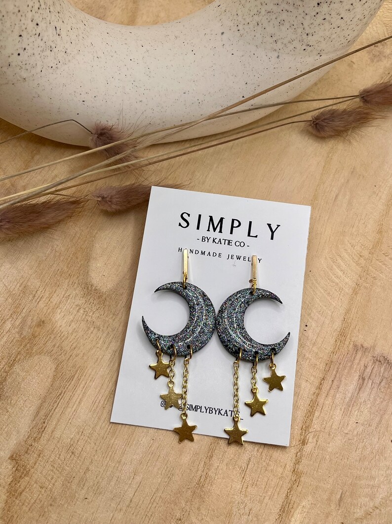 Op de afbeelding: Een paar oorbellen in de vorm van een halve maan met een donkere, glinsterende afwerking en bungelende goudkleurige sterbedels aan fijne kettinkjes, gepresenteerd op een witte kaart met de tekst SIMPLY BY KATIE CO. HANDMADE JEWELRY.