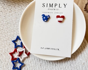 Op de afbeelding: Patriottische oorstekers, één blauw met witte sterren, de andere rood en wit. De oorbellen staan op een witte kaart met de tekst "SIMPLY - BY KATIE CO - HANDMADE JEWELRY".