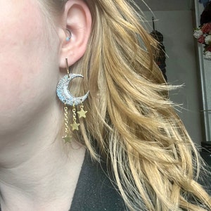 Op de afbeelding: Een oorbel in de vorm van een halve maan met een zilverkleurig glitteroppervlak en drie bungelende goudkleurige kettinkjes, elk eindigend in een kleine goudkleurige ster. De oorbel is bevestigd aan een dun verticaal staafje in de oorlel.