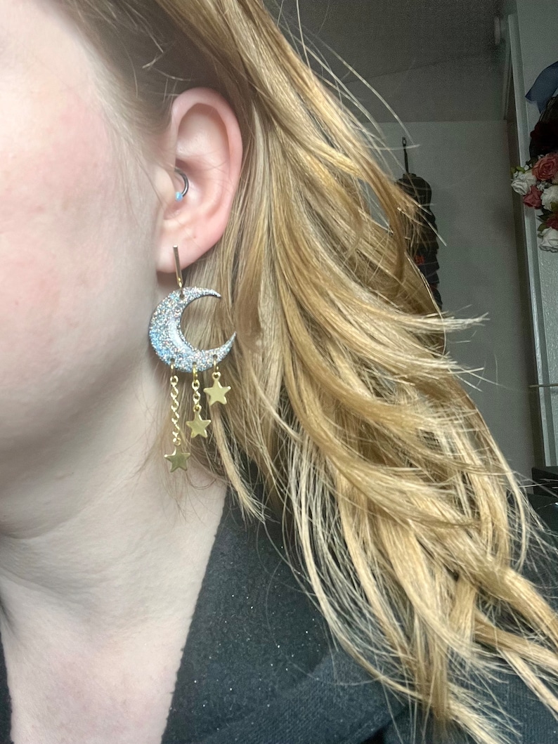 Op de afbeelding: Een oorbel in de vorm van een halve maan met een zilverkleurig glitteroppervlak en drie bungelende goudkleurige kettinkjes, elk eindigend in een kleine goudkleurige ster. De oorbel is bevestigd aan een dun verticaal staafje in de oorlel.