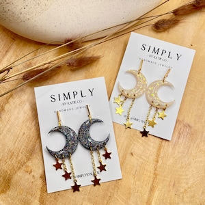 Op de afbeelding: Twee paar oorbellen in de vorm van een maansikkel op witte displaykaarten met het label SIMPLY BY KATIE CO. E&eacute;n paar heeft donkere glittermanen met bungelende donkere sterren aan gouden kettinkjes, het andere paar heeft iriserende witte manen.