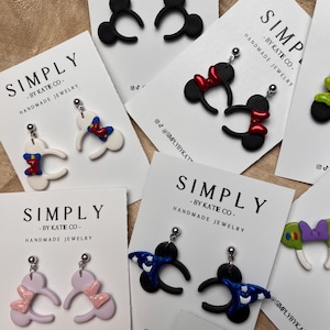 May include: A selection of handmade earrings in various designs, including Mickey Mouse ears with red bows, black ears, and other character-inspired styles. Each pair is presented on a white card with the text "SIMPLY - BY KATIE CO - HANDMADE JEWELRY".
