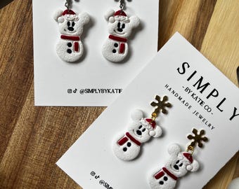 Op de afbeelding: Twee paar witte oorbellen in de vorm van een sneeuwpop met rode mutsen en sjaals, elk voorzien van een sneeuwvlokbedel. De oorbellen worden gepresenteerd op witte kaarten met SIMPLY BY KATE CO. HANDMADE JEWELRY en social media accounts.