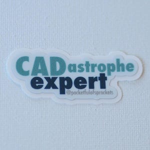 May include: A white sticker with the words "CADastrophe expert" in teal and navy blue, with the text "@pocketfulofsprockets" below. The sticker is on a white textured background.
