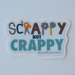Op de afbeelding: Een witte sticker met de tekst "SCRAPPY NOT CRAPPY" in verschillende kleuren en lettertypen. Het woord "SCRAPPY" bevat een schaar, een tandwiel en een liniaal in de letters. De sticker bevat ook de tekst "@pocketfulofsprockets".